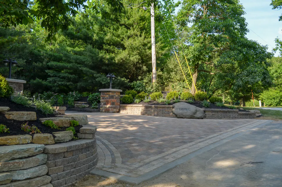 Paver driveway court with low walls, stone piers, and wooded planting
